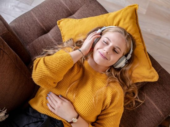 Blick von oben: Junge Frau mit Kopfhörer liegt auf der Couch und hört Musik, steht für Audio von FUNKE