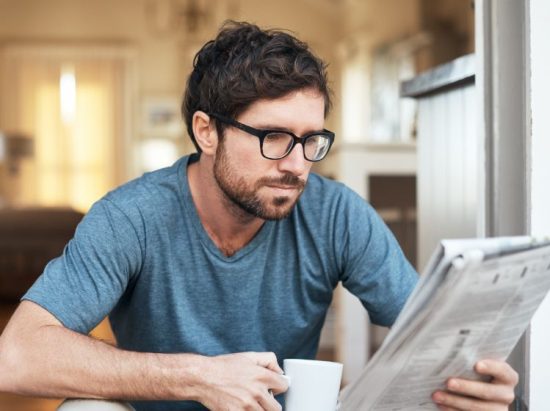 Junger Mann mit Brille und Kaffeebecher liest Zeitung und symbolisiert Anzeigenblätter
