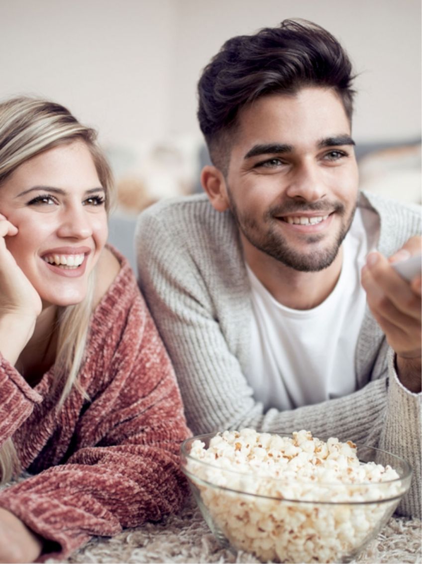 Next Level TV: Junges Paar liegt auf Teppich mit Schüssel Popcorn und Fernbedienung