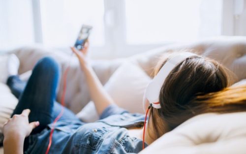Junge Frau liegt auf der Couch, trägt Kopfhörer und hört Radio auf Ihrem Smartphone, symbolisiert Online Radio (Audio)
